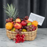 Canasta tejida llena de frutas frescas como piña, naranjas, manzanas, nectarinas, uvas y limones, decorada con lazo rojo y tarjeta de dedicatoria sobre mesa de mármol con fondo gris