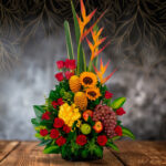 Arreglo floral vertical con heliconias, girasoles, rosas rojas, piñas decorativas, uvas y manzanas sobre mesa de madera con fondo oscuro