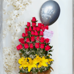 Arreglo de flores con varias filas de rosas rojas, lirios amarillos en la base, follaje verde, nube blanca decorativa y globo negro con texto Feliz Cumpleaños
