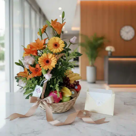 Cesta de regalo con flores frescas (lirios, gerberas, rosas) y frutas variadas, sobre mármol, con tarjeta y cinta.