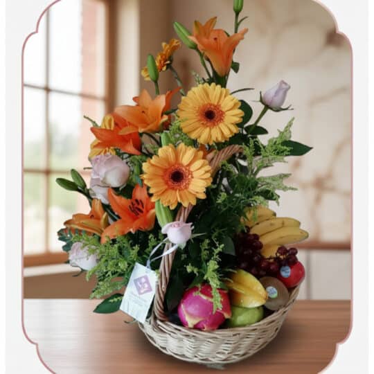 Cesta de mimbre con arreglo floral de rosas, lirios y gerberas, acompañada de frutas frescas como plátanos, uvas, manzanas y