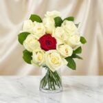 Arreglo floral CARLA con 12 rosas de tallo largo, una roja central y once blancas, en un jarrón de cristal sobre mármol.