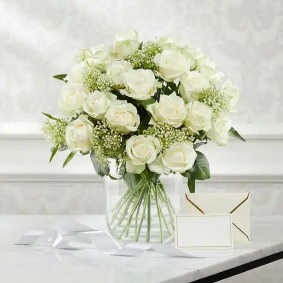 Arreglo floral Estefanía con 24 rosas blancas frescas en jarrón de cristal, tarjeta de regalo y cinta satinada sobre mármol.
