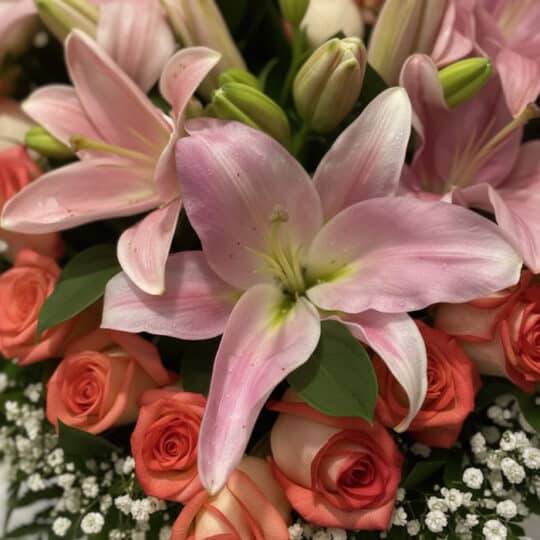 Primer plano extremo de un arreglo floral 'Feliz Contigo' con lirios rosados, rosas coral y pequeñas flores blancas, mostrand