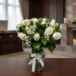 Arreglo fúnebre Pétalos del Alma con rosas blancas y follaje verde en un jarrón de cristal con lazo blanco, sobre una mesa de