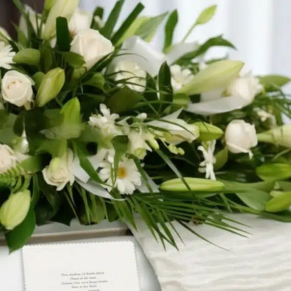 Arreglo fúnebre cubre caja con rosas blancas, lirios y follaje verde, con una cinta satinada blanca y una tarjeta de condolen