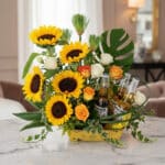 Arreglo floral vibrante con girasoles, rosas blancas y naranjas, follaje verde y tres botellas de cerveza Corona, ideal para