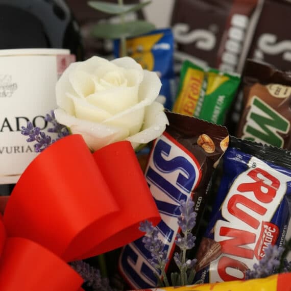 Primer plano extremo de una rosa blanca con gotas de rocío, lavanda, lazo rojo, chocolates Snickers y Run, y botella de vino.
