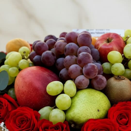 Primer plano macro de la Caja Armani, mostrando uvas rojas y verdes, manzanas, pera, kiwi y rosas rojas con gotas de rocío.