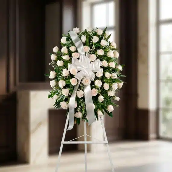 Corona Fúnebre Serenidad: Elegancia y Respeto con Rosas Blancas Corona fúnebre Serenidad con rosas blancas y follaje verde, lazo blanco, sobre un trípode en una sala de velación premium con