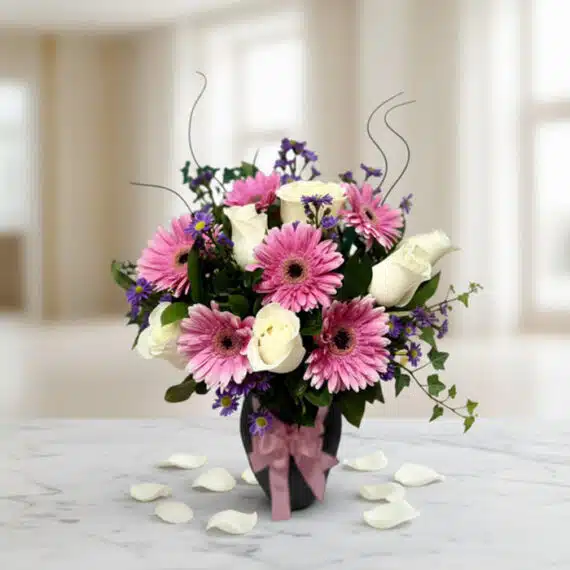 Jarrón Primavera: Elegante Arreglo Floral con Rosas y Gerberas Jarrón Primavera con un elegante arreglo floral de rosas blancas, gerberas rosas y pequeñas flores moradas, sobre una mesa de