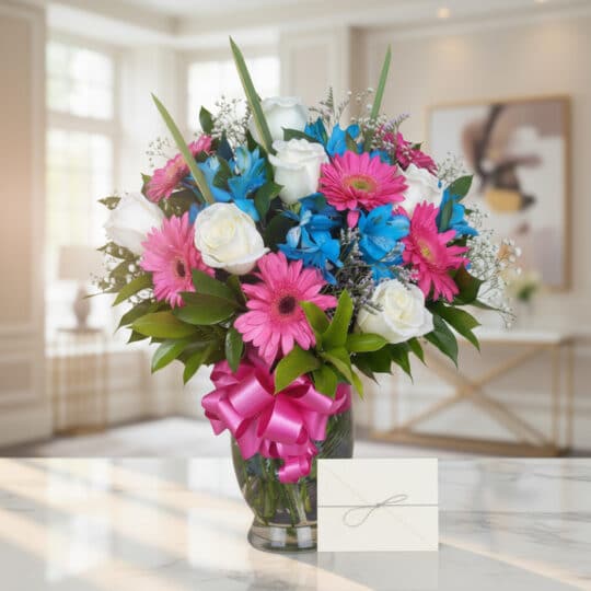 Jarrón Primavera con rosas blancas, gerberas fucsia y alstroemerias azules, sobre una mesa de mármol en un ambiente luminoso