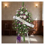 Pedestal fúnebre Esteban con arreglo floral de lirios blancos, rosas moradas y follaje verde, banda blanca con nombre 'Esteba